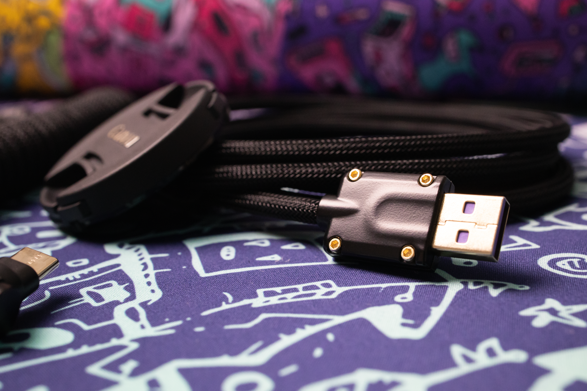 Closeup of CNC USB A connector with purple PCB and wrapped black on black sleeved Teleios mechanical keyboard cable with a Canon camera lens out of focus on a multi colored desk mat. 